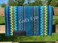 Hand-woven western show pad with a rich teal blue base featuring geometric diamond patterns in lime green, bright yellow, royal blue, turquoise, and rust orange accents with black wear leathers. 